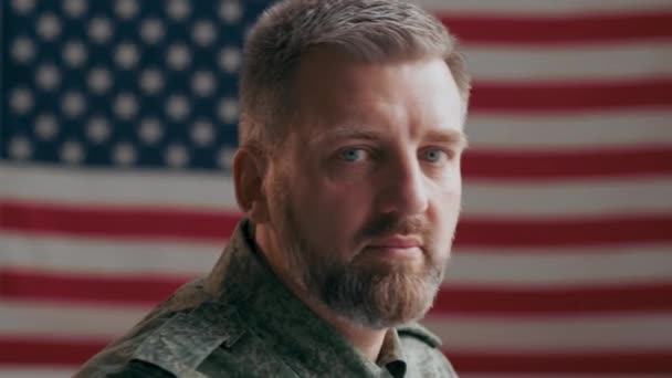 Porträt eines ernsthaften männlichen Offiziers mittleren Alters, der sich dreht und in die Kamera schaut, während er vor der US-Flagge posiert - Filmmaterial, Video