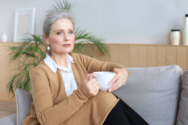 Happy mature woman resting on comfortable sofa drink coffee or tea, looking away, relaxing on cozy couch at home enjoy hot beverage - Photo, image