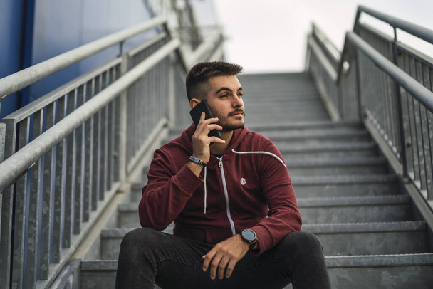 A shallow focus of a young male with a beard sitting on the steps outdoors and talking on a phone - Foto, Bild