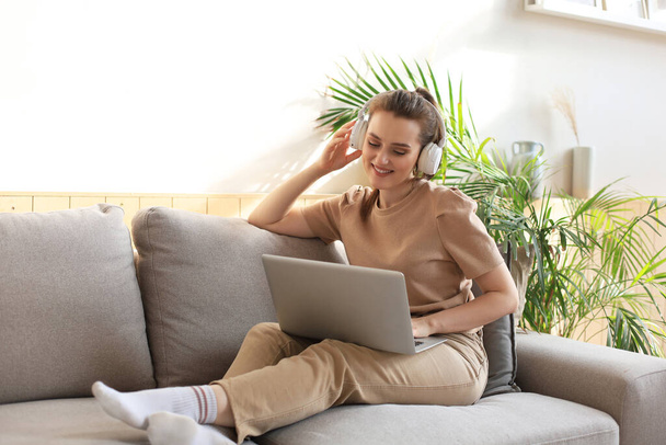 Smiling young woman with headphones and laptop on the sofa - Фото, изображение