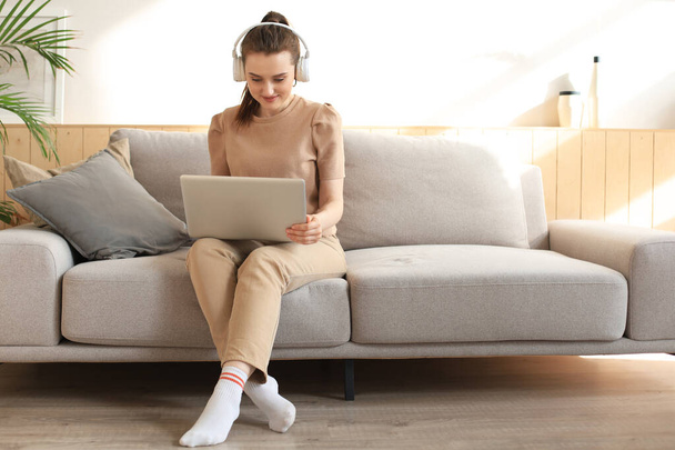 Smiling young woman with headphones and laptop on the sofa - Foto, afbeelding