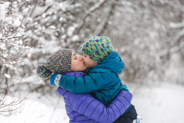 Дитина обіймає маму. Хлопчик з матір'ю на зимовій прогулянці. Дитина в плетеному капелюсі цілує свою матір. Жінка підтримує свого сина. Матері. Сім'я проводить разом час. Сімейні цінності. - Фото, зображення