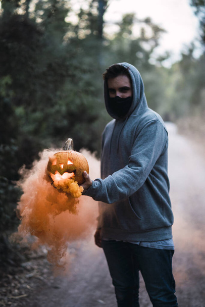 A vertical shot of a man holding a carved pumpkin with an orange smoke - the concept of Halloween - Foto, Imagen