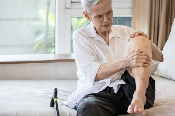 Wyczerpana azjatycka starsza kobieta trzyma kolano z bólem w stawie kolanowym, pacjent w podeszłym wieku masuje kolana dłońmi, bolesna ekspresja twarzy, choroba reumatyczna, zapalenie kości i stawów u osób starszych - Zdjęcie, obraz