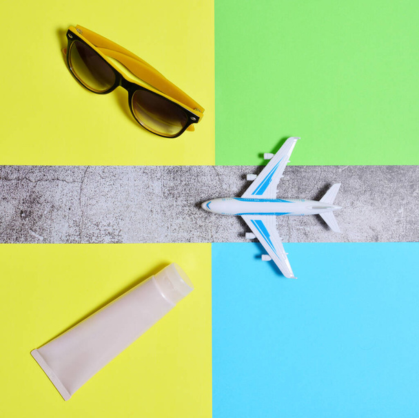 Podyumdaki oyuncak uçak, güneş kremi, güneş gözlüğü. Tatil uçuşu konsepti, sıcak bölgelerde tatil. Sarı yeşil ve mavi arka plan. düz alan, kopyala boşluk. - Fotoğraf, Görsel