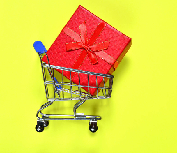 klein winkelwagentje op wielen op een gele achtergrond, met een rood cadeau. shopping concept, verjaardagen, kerst en nieuwjaar, kortingen in winkelcentra. kopieerruimte. - Foto, afbeelding