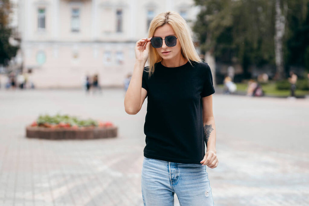 Elegante chica rubia con camiseta negra y gafas posando contra la calle, estilo de ropa urbana. Fotografía callejera - Foto, Imagen