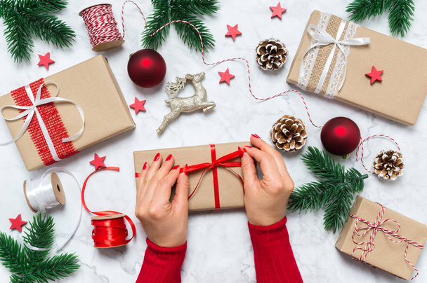 Female hands make bow on Christmas present. Festive holiday composition with woman hands, gift boxes, pine tree, cones, baubles and decoration on marble background. Holiday theme. Top view, flat lay. - Photo, Image