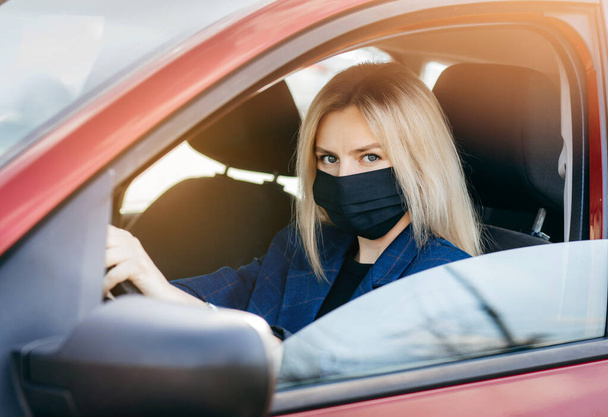 Νεαρό κορίτσι με μαύρη μάσκα κάθεται σε ένα αυτοκίνητο, προστατευτική μάσκα κατά του κορωναϊού, οδηγός σε ένα δρόμο της πόλης κατά τη διάρκεια μιας επιδημίας του κορωναϊού - Φωτογραφία, εικόνα