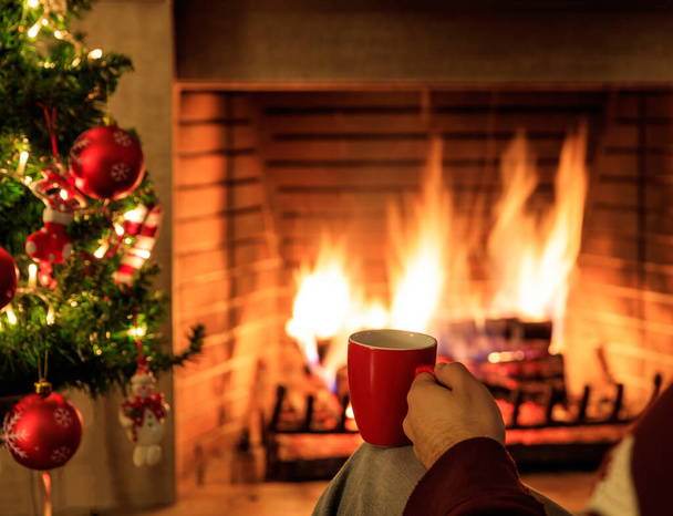 Man holding a red cup of coffee or tea on burning fireplace background. Warm cozy home interior, winter time - Photo, Image