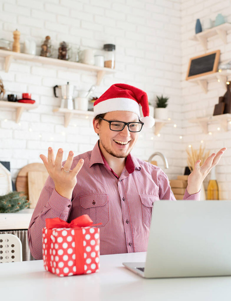 Karácsonyi online üdvözlet. Boldog fiatalember télapó kalapban üdvözöli barátait video chat vagy hívja a laptop ül a konyhában - Fotó, kép