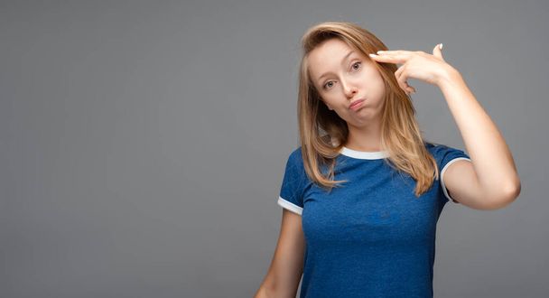 Funny young blonde female shoots in temple, tilts head, dressed in casual blue t shirt, demonstrates suicide gesture, isolated on gray background with blank space - Photo, image