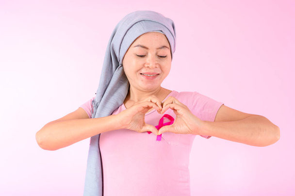 Muslim woman wearing a hijab with a ribbon on her chest shows prevention of breast cancer. On a pink backdrop, breast cancer concept, cancer prevention concept. - Foto, Imagen