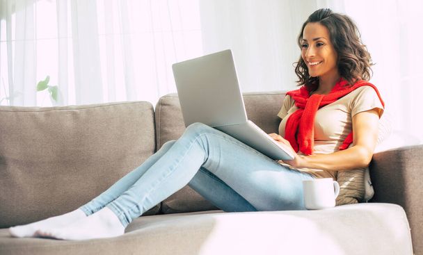 Happy cute brunette woman is relaxing on the couch at home and using laptop - Фото, изображение