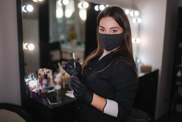 Portrait of professional female brow master in black robe with black gloves and black protective mask use brush and henna for eyebrows - Photo, image