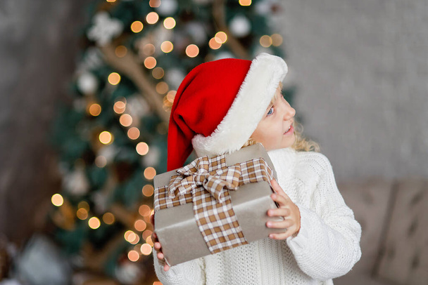 Happy excited girl child holding christmas gift box. Child giving a Christmas present in decorated room. christmas and people concept. New Year Happy holidays. kid satisfied with present. - Φωτογραφία, εικόνα
