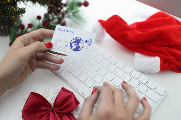 Mãos femininas segurando cartão de crédito bancário perto de teclado e chapéu de santa claus e árvore de natal. - Foto, Imagem