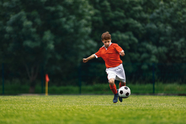 Soccer shooting. Boy kicking soccer ball on grass field. Young football player in action running jumping, and shooting the ball. Junior level sports competition. Footballer in red jersey shirt - Photo, image