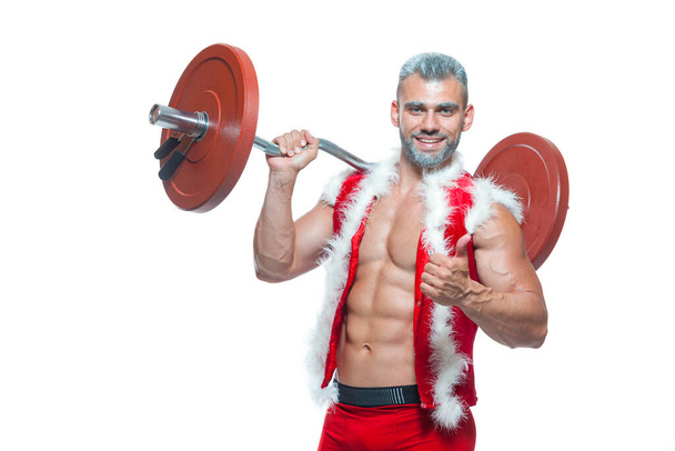 Sexy Santa Claus. Bodybuilder young handsome santa clause smile holds a barbell with red pancakes on the shoulder and shows off abs cubes at New Years eve and Christmas winter holiday white background - Foto, Imagen