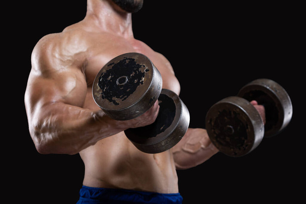 Close up of muscular sportsman lifting heavy dumbbells showing his strong biceps isolated on black backgrounds - Foto, afbeelding