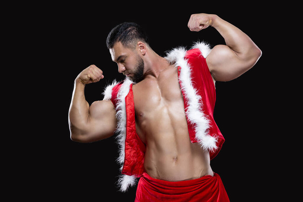 Young sexy Santa Claus in a red carnival costume on a naked body shows off his strong biceps isolated on black background - Foto, imagen