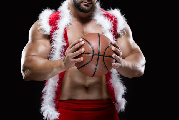Front view portrait of an attractive muscular Santa Claus holding a basketball with both hands isolated on black background - Foto, immagini