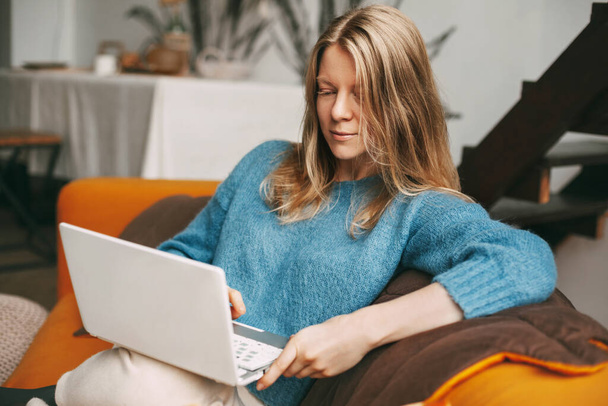 Mujer joven utiliza el ordenador portátil, comprueba las noticias de correo electrónico en línea sentado en el sofá. Hermosa chica trabajando en una computadora, viendo un webinar, estudiando en casa - Foto, imagen
