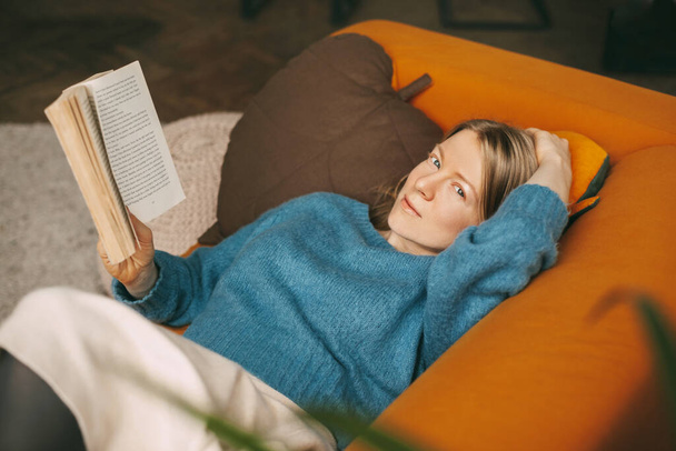 Una hermosa chica se está relajando en el sofá de la sala de estar, leyendo su novela favorita. Linda y pensativa chica está tumbada en el sofá y leyendo un libro. Trabajo en línea, freelance, distancia social - Foto, imagen