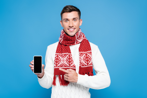 Усміхнений молодий дорослий чоловік, одягнений светр і шарф, показує і вказує пальцем на смартфон ізольовано на синьому
 - Фото, зображення