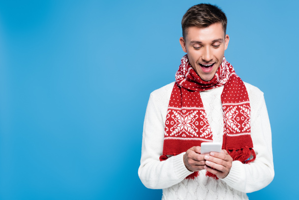 Emocionado hombre con bufanda de punto y suéter, mensajes de texto en el teléfono inteligente aislado en azul - Foto, Imagen