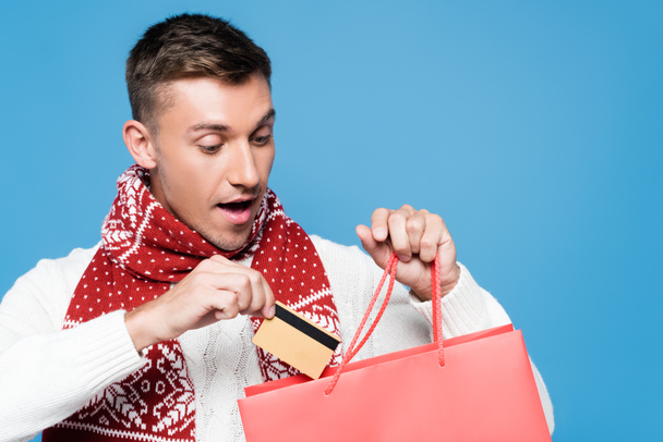 Portret zaskoczonego młodego mężczyzny, wkładającego kartę kredytową do czerwonej papierowej torby odizolowanej na niebiesko - Zdjęcie, obraz