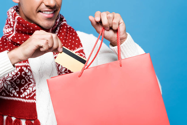 Cropped view of smiling man putting credit card in red paper bag isolated on blue - Zdjęcie, obraz
