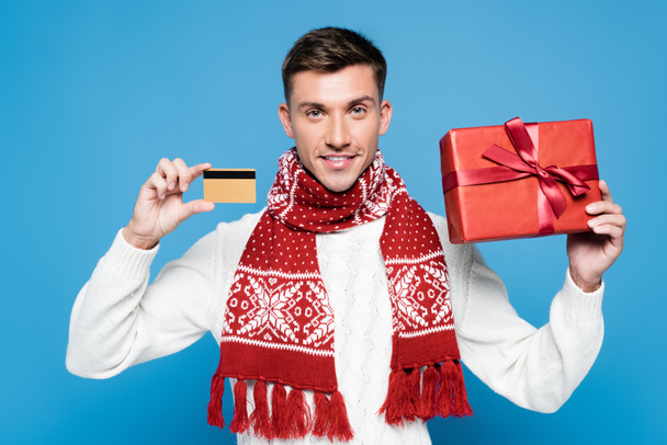Hombre sonriente con bufanda y suéter, mostrando regalo envuelto y tarjeta de crédito aislada en azul - Foto, imagen