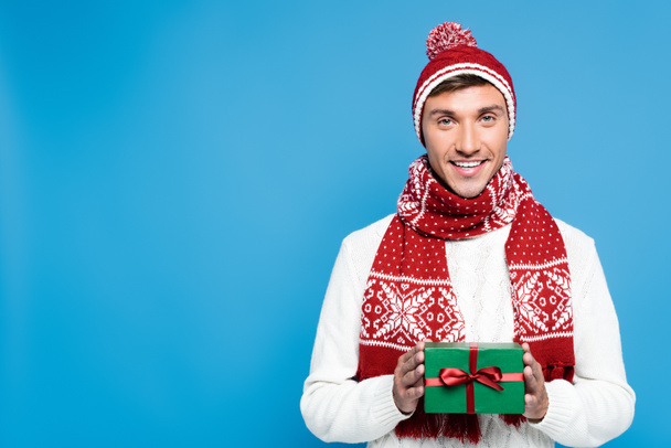 Glimlachende jonge volwassen man in warme kleding houden geschenkdoos terwijl kijken naar camera geïsoleerd op blauw - Foto, afbeelding