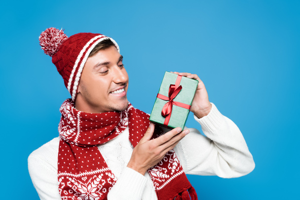 Hombre feliz en sombrero de punto con bola que muestra una pequeña caja de regalo aislada en azul - Foto, imagen