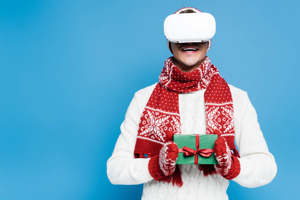 Χαμογελώντας άνθρωπος με ζεστά ρούχα, χρησιμοποιώντας ακουστικά VR και κρατώντας μικρό κουτί δώρου απομονώνονται σε μπλε - Φωτογραφία, εικόνα