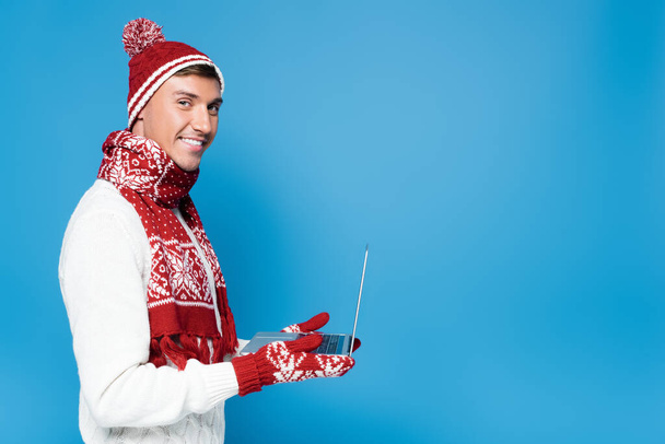 Happy man in warm clothing, holding laptop isolated on blue - Foto, imagen