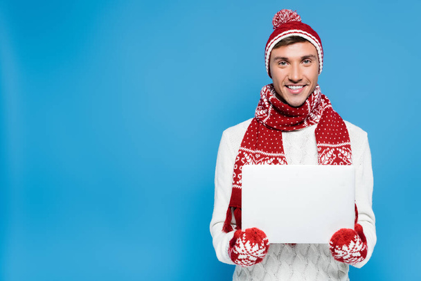Front view of smiling young adult man in warm clothing, standing with laptop isolated on blue - Zdjęcie, obraz
