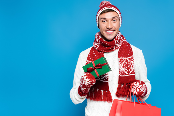 Sorrindo jovem adulto homem em roupas quentes segurando pequena caixa de presente e saco de papel vermelho isolado em azul - Foto, Imagem
