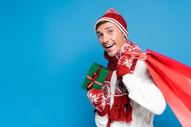 Excited young adult man with red paper bags behind back, holding small wrapped present isolated on blue - Foto, Imagen