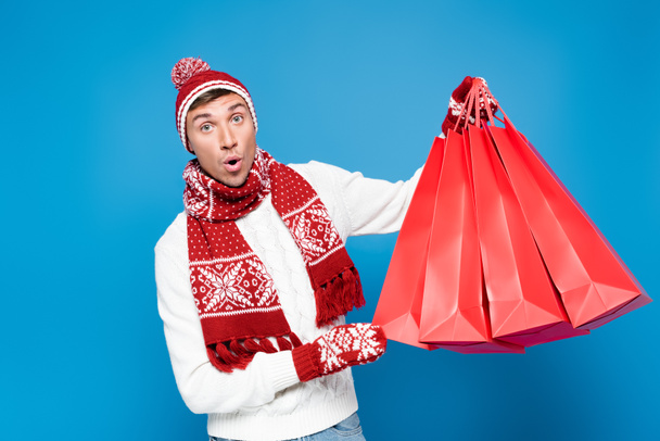 Shocked man in mittens lifting red paper bags, while looking at camera isolated on blue - Photo, Image