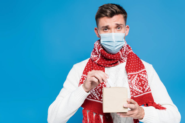 Giovane uomo adulto in maschera medica contenente scatola di tessuti isolati su blu - Foto, immagini