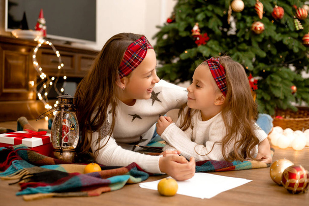Due ragazze sul pavimento di legno della casa accanto all'albero di Natale che scrivono una lettera a Babbo Natale facendo auguri - Foto, immagini