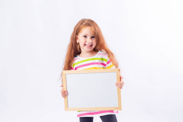 kleines Mädchen mit langen roten Haaren lächelt und hält ein weißes Reißbrett auf weißem Hintergrund. Raum für Text. - Foto, Bild