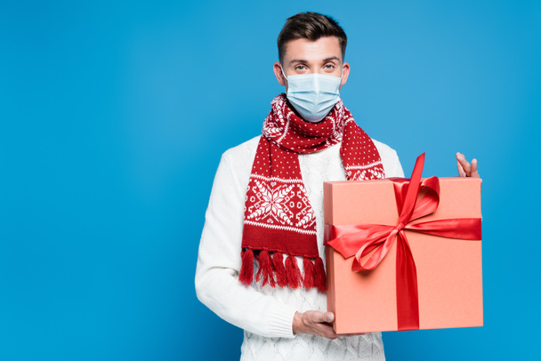 Junger erwachsener Mann in medizinischer Maske hält Geschenkbox in der Hand und blickt isoliert in die Kamera auf blauem Grund - Foto, Bild