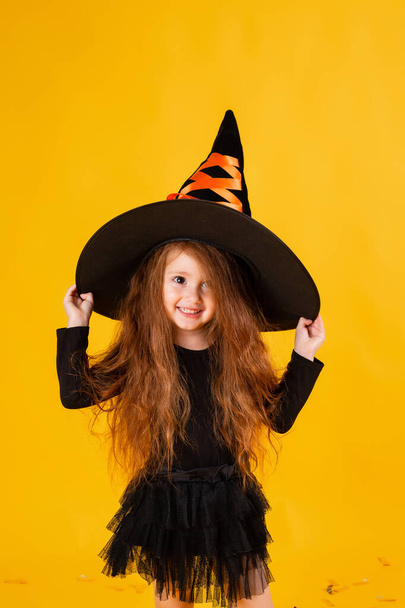 little girl with long red hair  in a Halloween witch costume on a yellow background. Halloween for children, space for text - Foto, Imagen