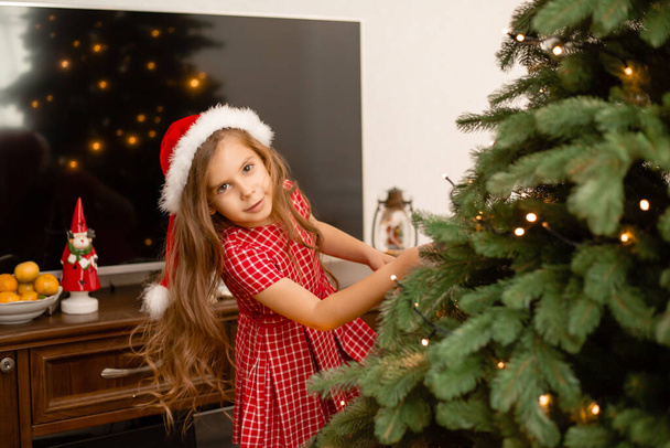 Schattig klein meisje in rode jurk versieren kerstboom thuis - Foto, afbeelding