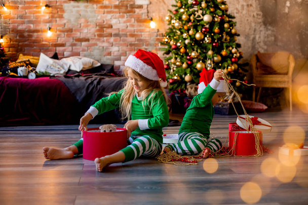 Δύο μικρά παιδιά με πιτζάμες και χριστουγεννιάτικα καπέλα ξεπακετάρουν χριστουγεννιάτικα δώρα στο δωμάτιο - Φωτογραφία, εικόνα