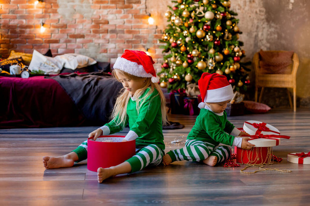 Twee jonge kinderen in pyjama 's en kerstmutsen pakken kerstcadeaus uit in de kamer - Foto, afbeelding