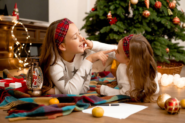Δύο κορίτσια στο ξύλινο πάτωμα του σπιτιού δίπλα στο χριστουγεννιάτικο δέντρο γράφοντας ένα γράμμα στον Άγιο Βασίλη κάνοντας ευχές - Φωτογραφία, εικόνα